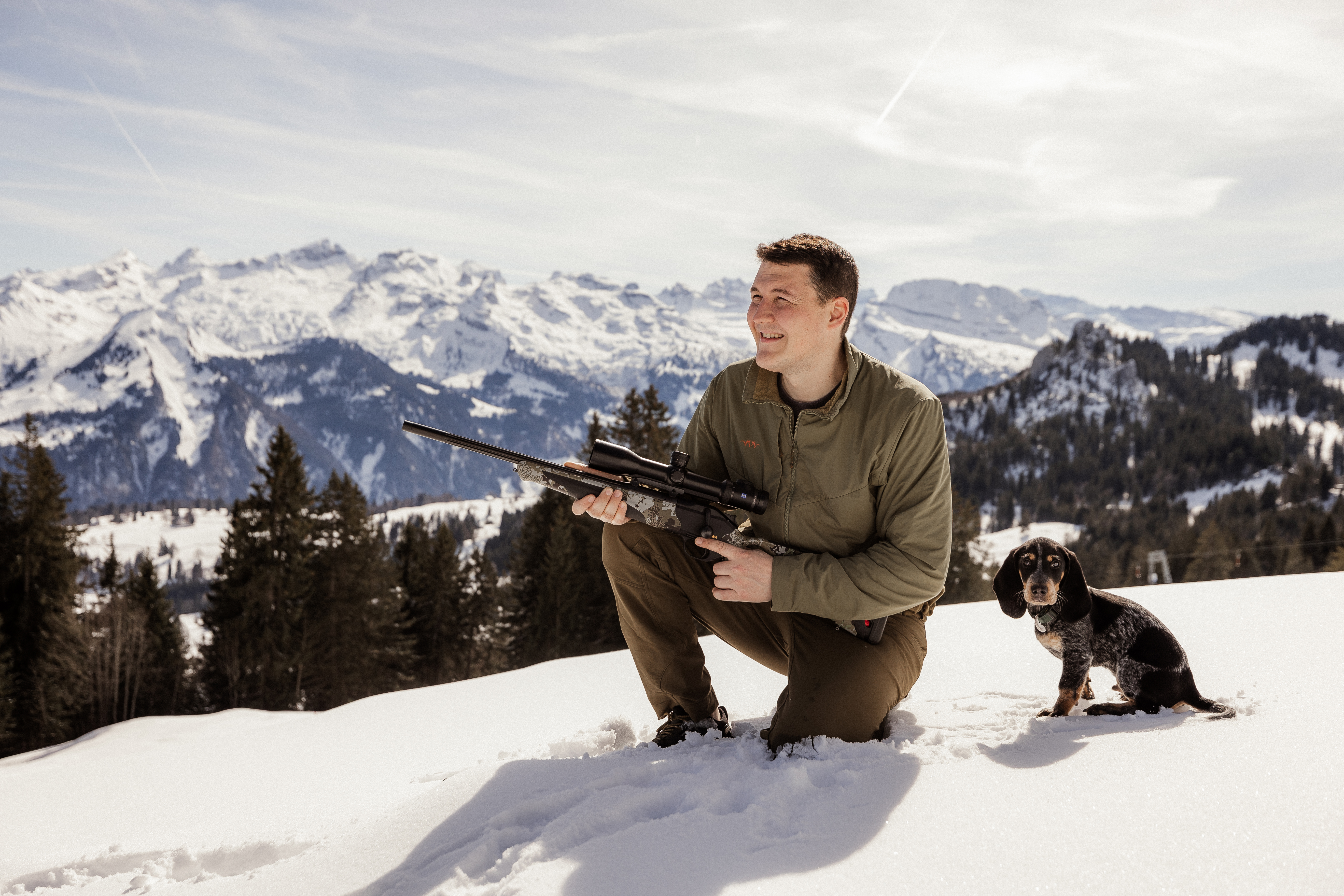 Man mit Gewehr hockt neben Hund auf schneebedeckten Berg